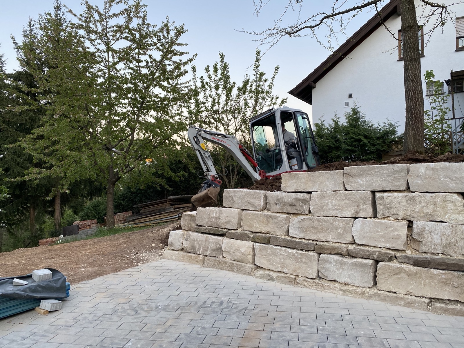 Fertig gesetzte Natursteinmauer aus großen hellen Blöcken neben einer frisch gepflasterten Fläche, dahinter der Minibagger.