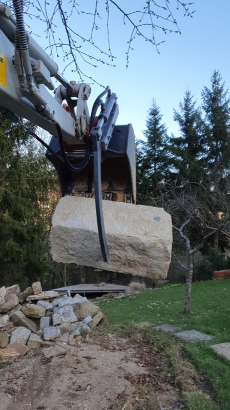 Der Bagger hebt mit Greifer und Schaufel einen großen Sandsteinblock an.