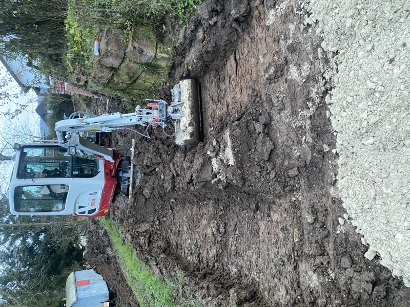 Ausgehobene Trasse für einen Weg entlang einer alten Steinmauer, vorne liegt Schotter bereit.
